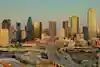Aerial photograph of downtown Dallas, Texas, captured during golden hour with warm sunlight illuminating the city's modern skyscrapers and urban infrastructure. The image features the central business district with its distinctive mix of glass towers and concrete high-rises, including prominent buildings that define the Dallas skyline. In the foreground, the complex network of elevated highways and interchanges creates geometric patterns as they weave through the urban landscape, connecting different parts of the metropolitan area. The photograph captures the contrast between the towering financial district and the lower-rise buildings in the surrounding neighborhoods, with clear skies providing excellent visibility across the sprawling Texas metropolis. • Authorization was obtained from the FAA for this operation in restricted airspace.